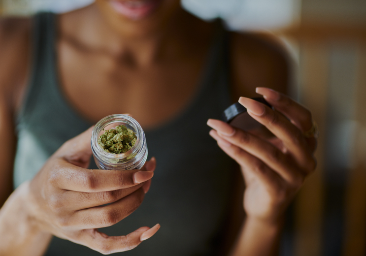 A close-up of a woman holding a small glass jar filled with cannabis buds. She is unscrewing the black lid while examining the contents. Her nails are long and painted in a neutral shade, and she is wearing a dark green tank top. The background is softly blurred, suggesting an indoor setting with warm lighting. 
