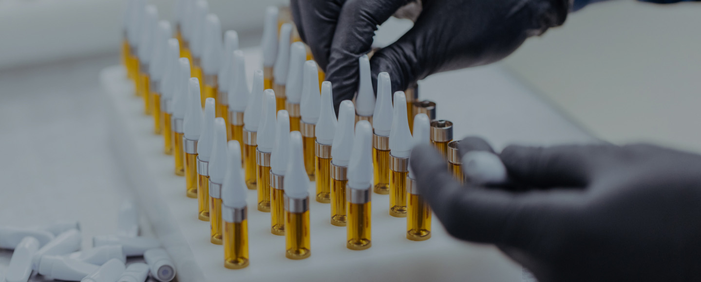 Close-up of a technician wearing black gloves assembling vape cartridges filled with golden cannabis oil in a laboratory setting.